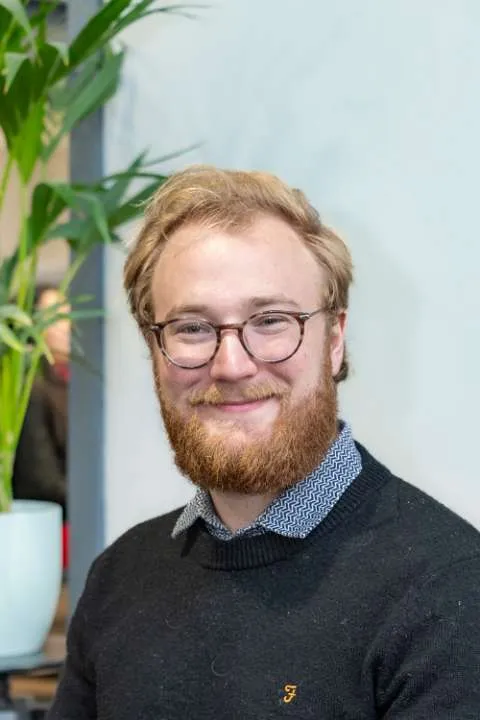 A portrait of a blonde man, wearing a blue jumper with a collared patterned shirt